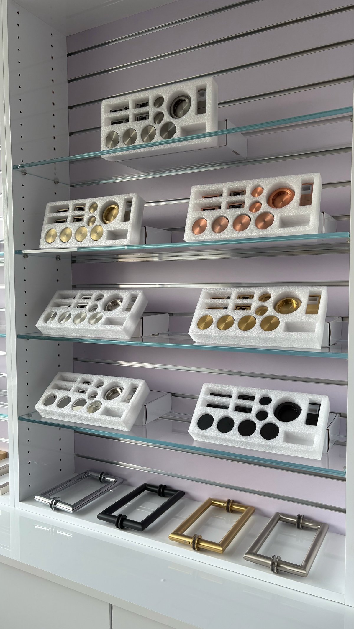 Metal shelving unit displaying white trays containing various decorative hardware items including knobs, hinges, and fixtures in different finishes